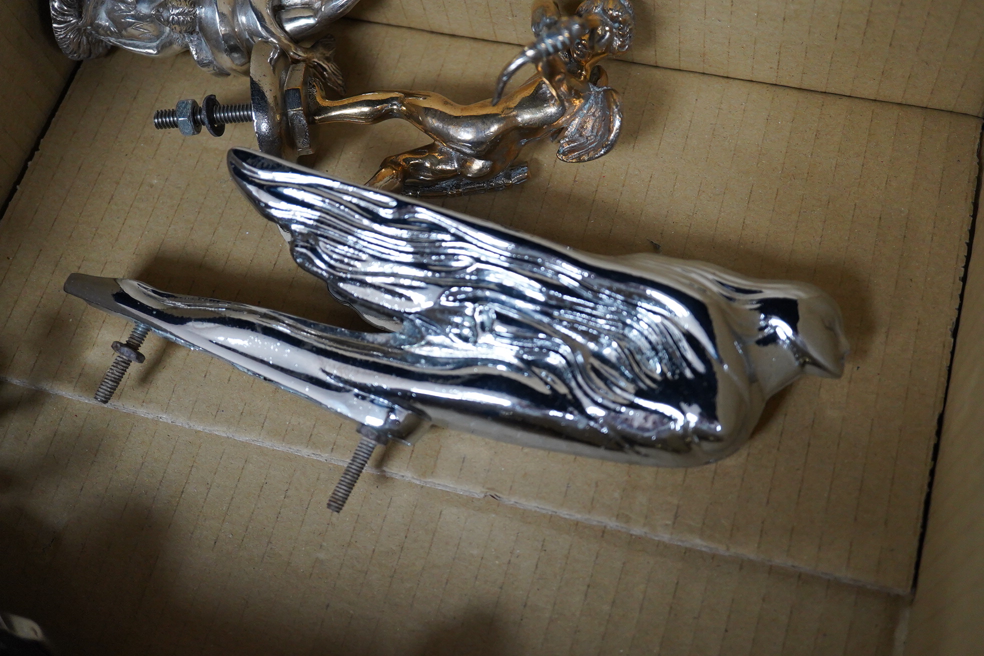 Six chrome and brass car mascots, including; a figure with a dolphin, a figure of Cupid, a grotesque in the form of a devil, a figure of a Tudor gentleman, an Art Deco figure of a flying woman, etc.